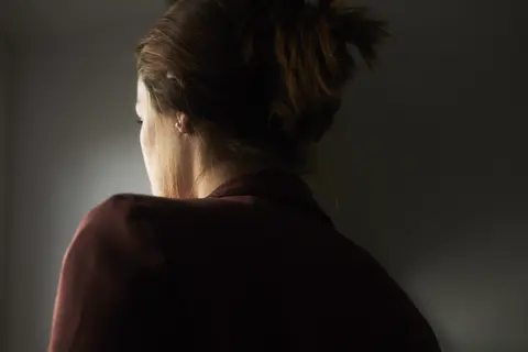 Frau von hinten im dunklen Oberteil, seitlich von warmem Licht beleuchtet, vor schlichtem dunklem Hintergrund.