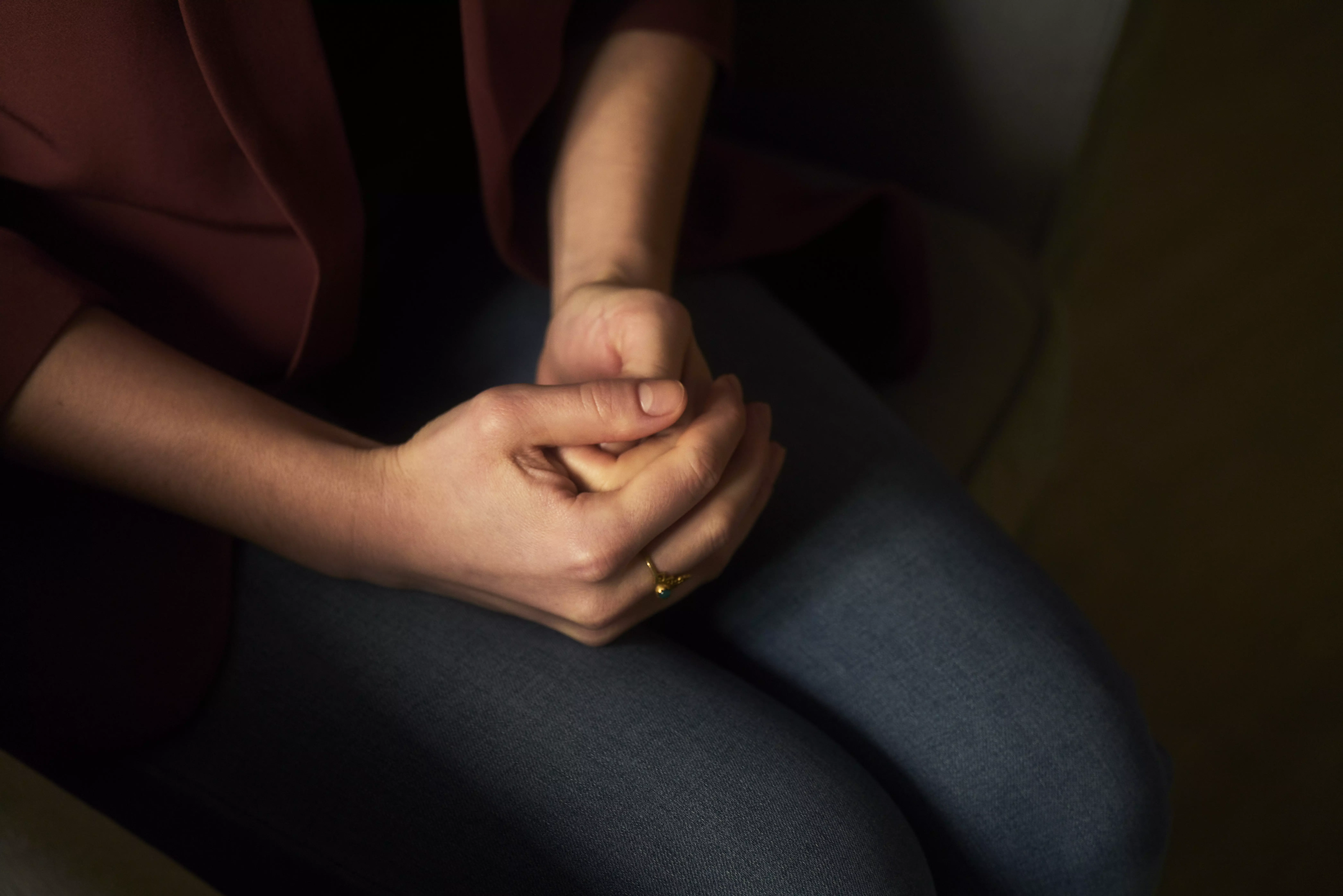 Nahaufnahme einer sitzenden Person mit gefalteten Händen im Schoß, rotem Oberteil und blauer Jeans, warm und gedämpft beleuchtet.