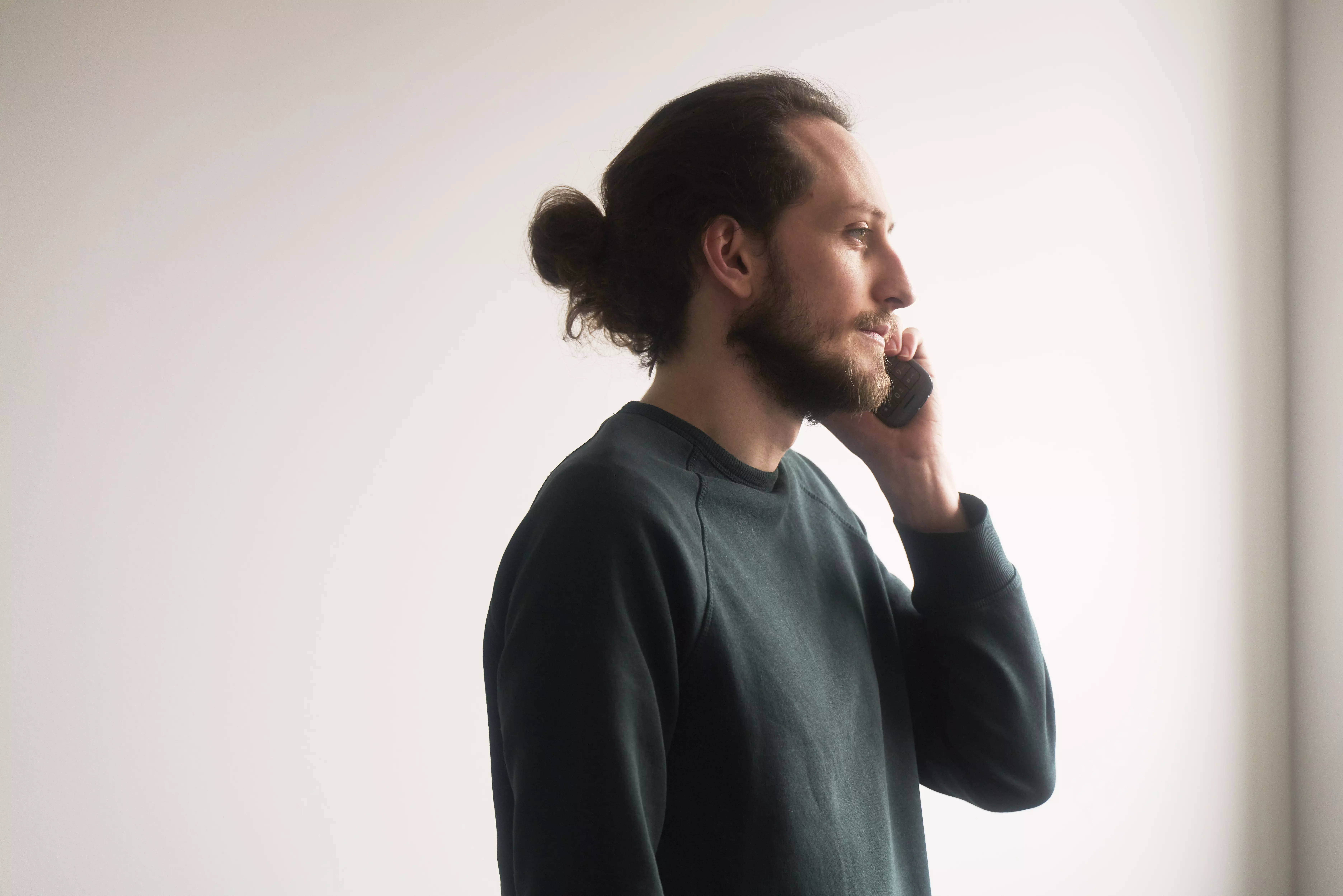 Seitlich stehender Mann mit dunklem Dutt telefoniert in einem hellen, minimalistischen Innenraum; weiches Tageslicht, ruhige Stimmung.